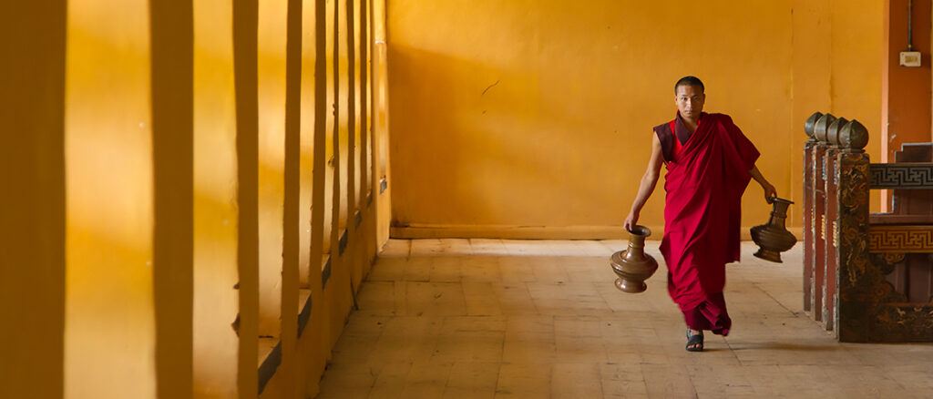 Monk in red robe carries copper colored urns down hallway in golden light